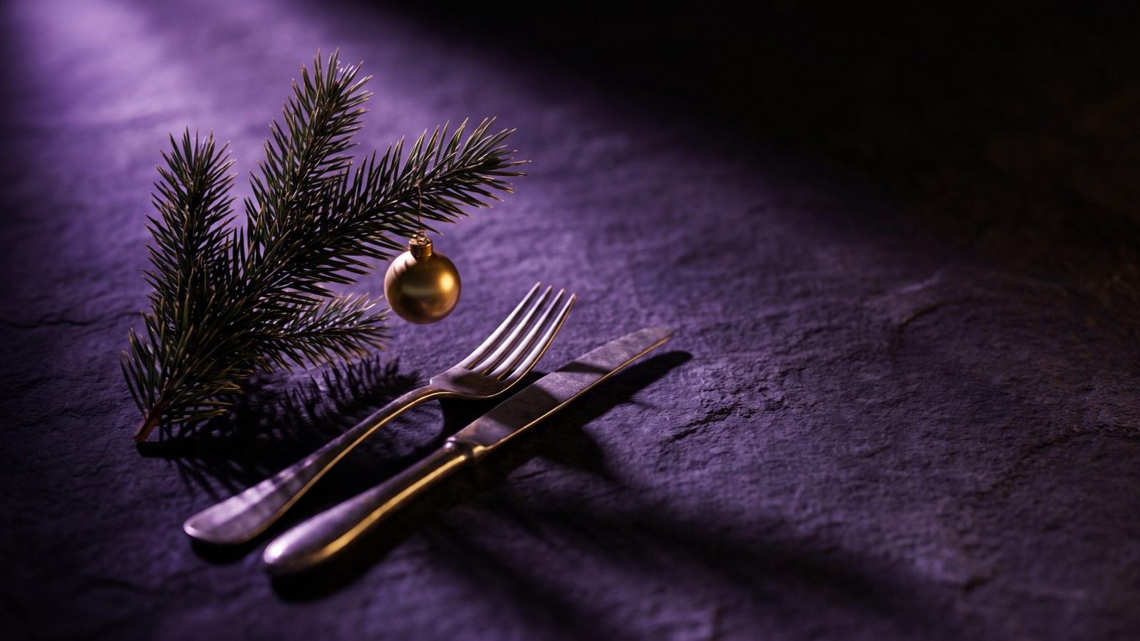 Ein Tannenzweig mit einer goldenen Kugel liegt neben Messer und Gabel auf dunklem Stein